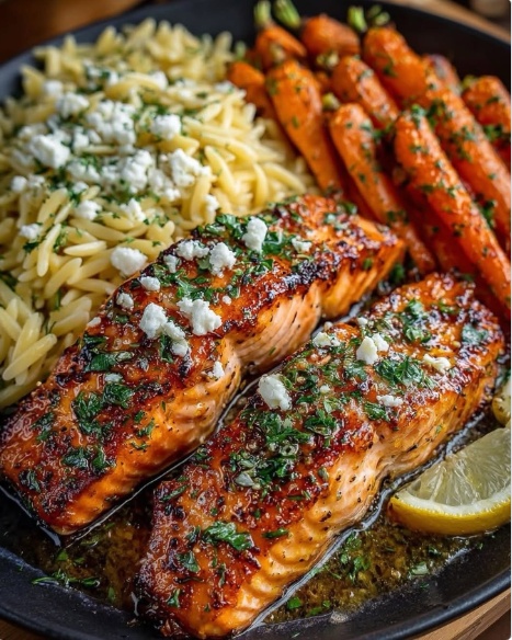 A plate featuring two glazed honey garlic salmon fillets garnished with fresh parsley and feta cheese, served with orzo and roasted baby carrots.