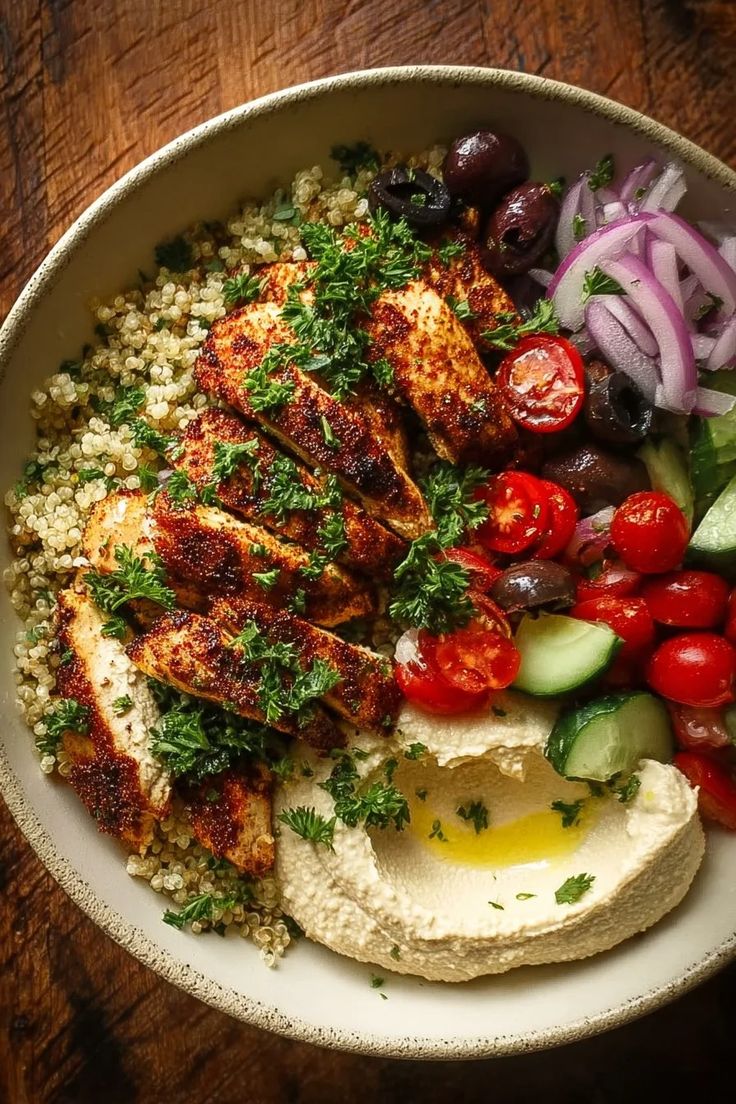 A loaded Mediterranean Chicken Quinoa Bowl featuring spiced grilled chicken, quinoa, sliced tomatoes, cucumbers, and a side of smooth hummus.