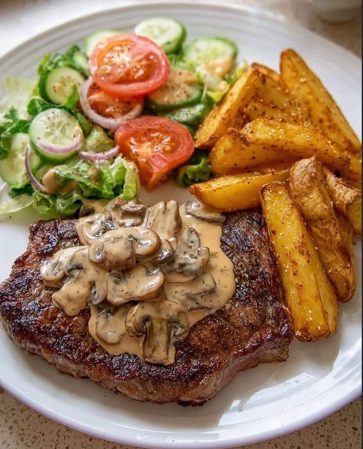A pan-seared juicy steak topped with creamy mushroom sauce, served with golden potato wedges and a fresh garden side salad on a white plate.